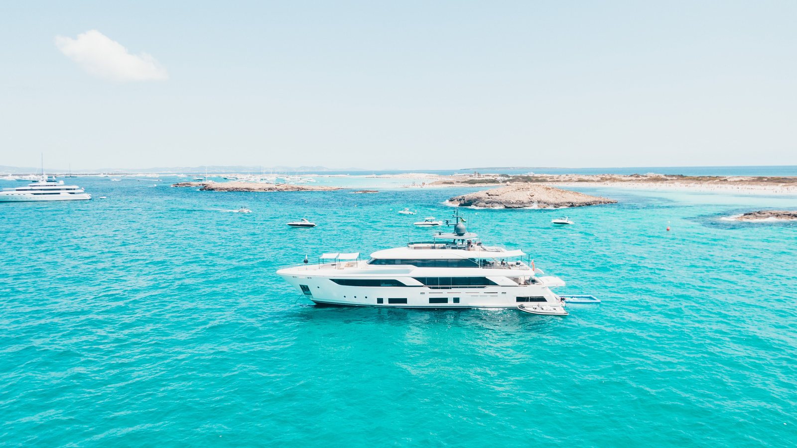 Aerial view of a superyacht at anchor in turquoise water with a tender alongside, beach and rocks behind