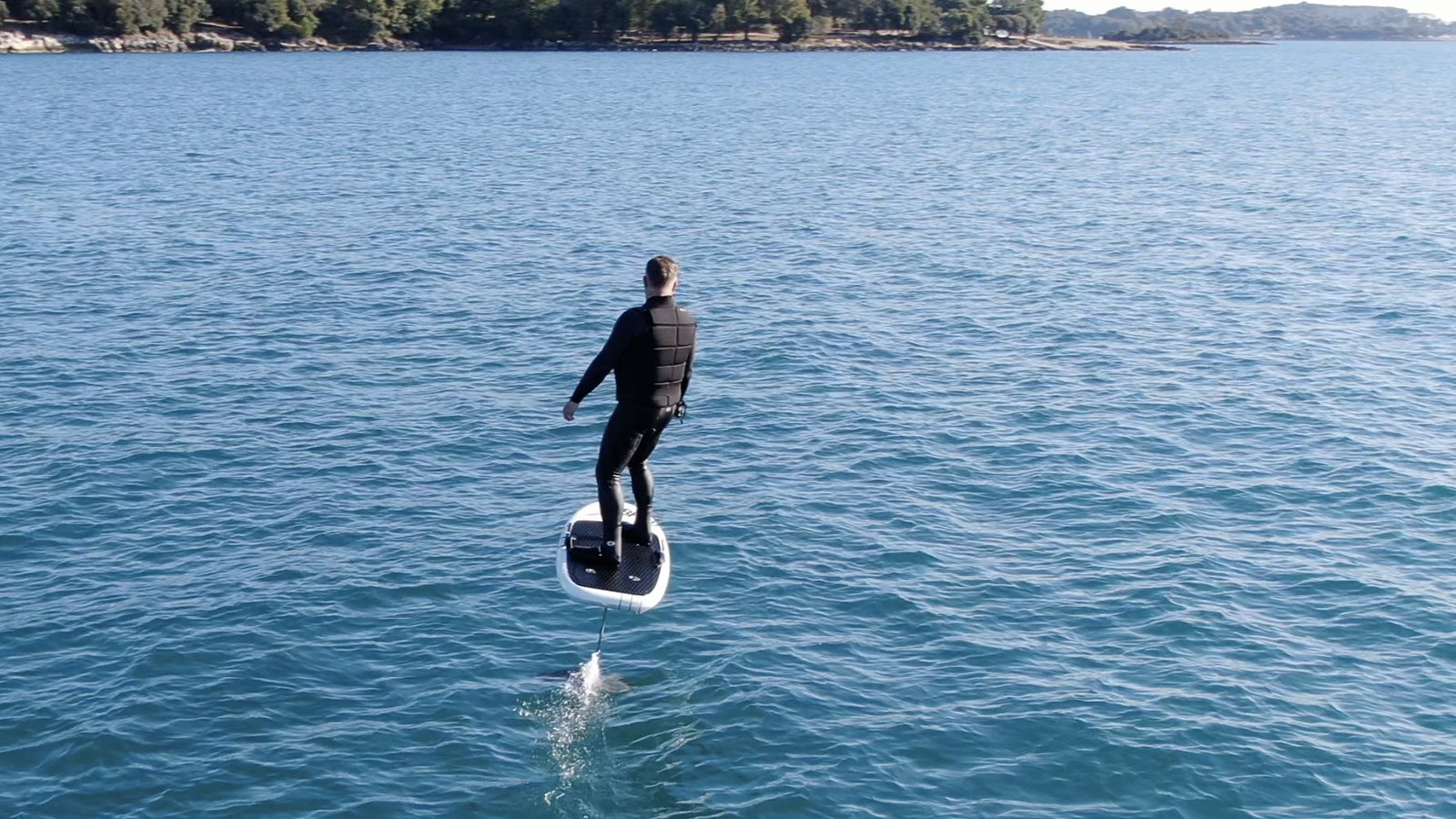 Drone view of a rider in a wetsuit and impact vest flying on an eFoil over clear blue water, wooded coast behind