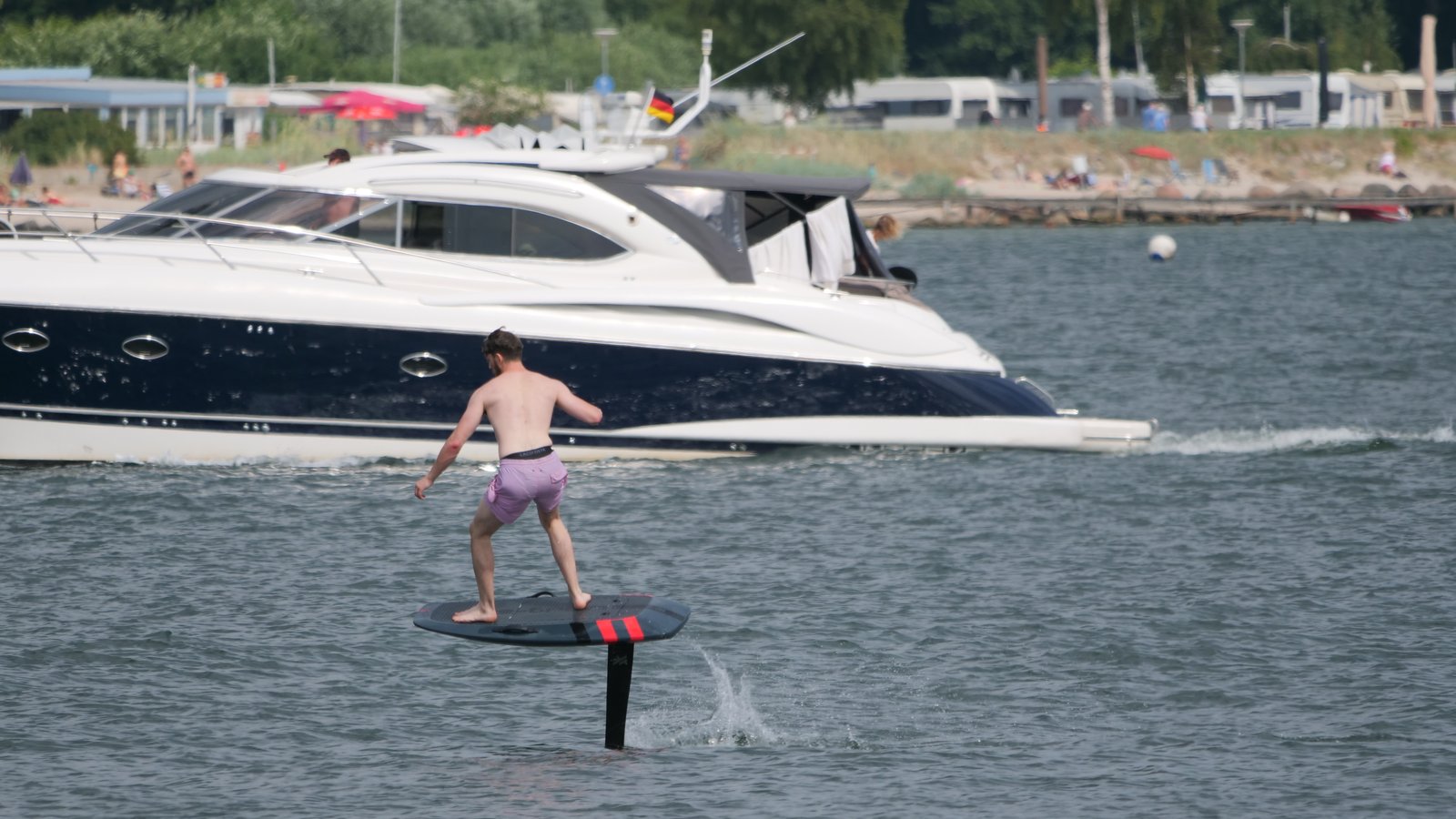 A rider on an eFoil flying past a motor yacht at sea, beach in the background