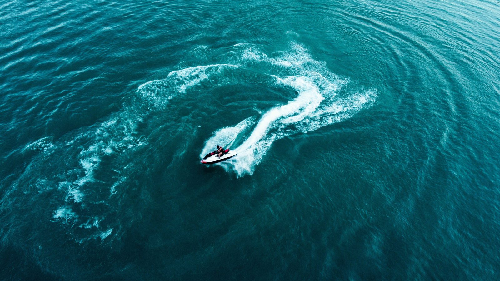 Aerial view of a jet ski carving a curved white wake through turquoise sea