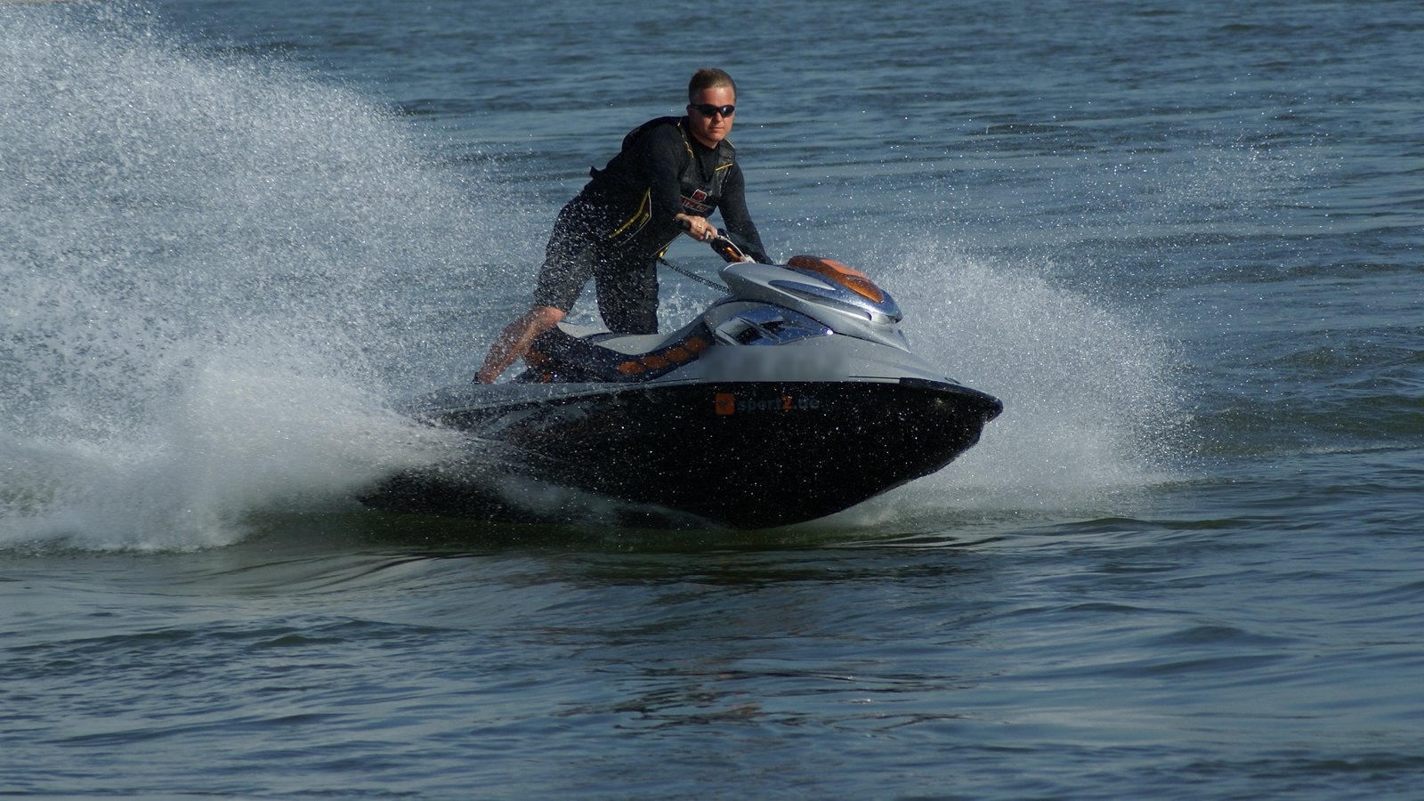 A rider in sunglasses and a buoyancy vest carving a jet ski through a tight turn with a wall of spray behind
