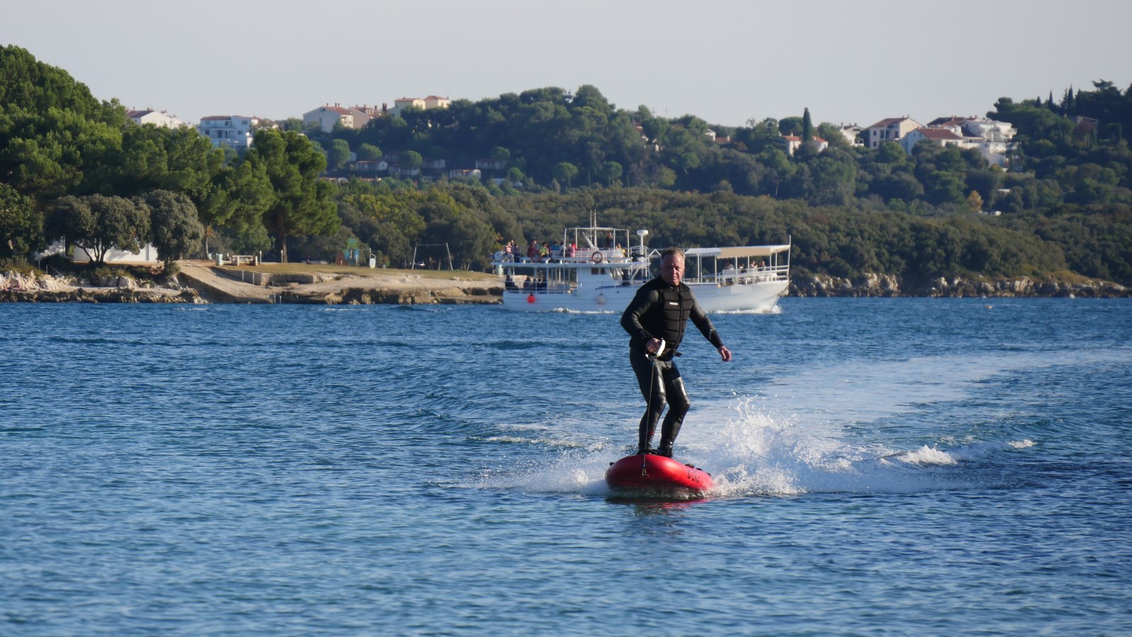 A rider in a wetsuit and impact vest on a red electric jetboard throwing spray in a bay, an excursion boat and wooded coast behind