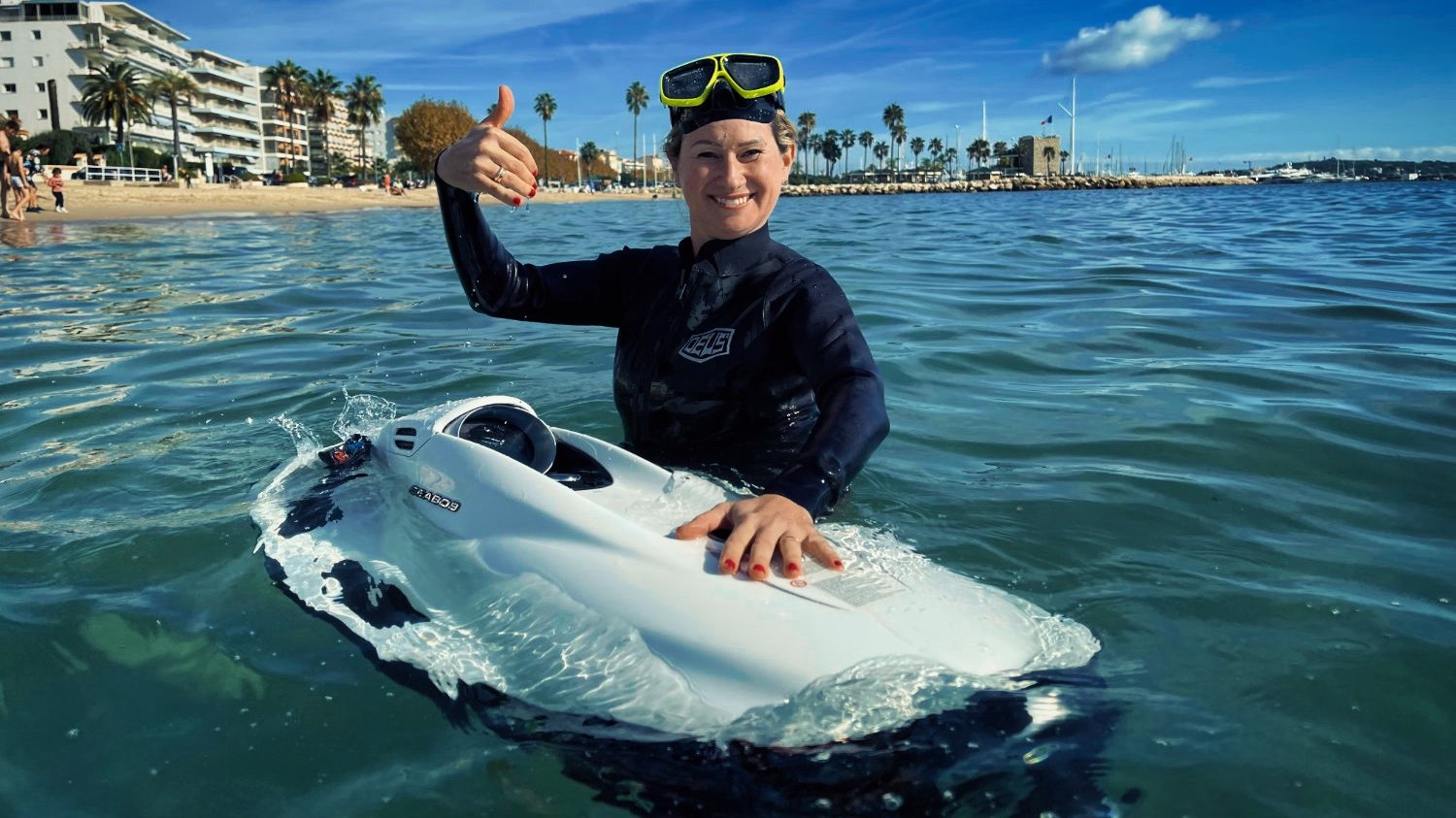 A smiling rider in a wetsuit and diving mask with a white SEABOB in clear sea water, beach and palm trees behind