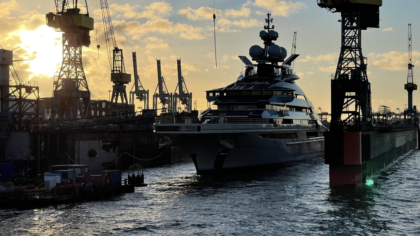 A large superyacht in a dry dock between shipyard cranes at sunset